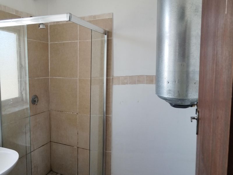 A functional bathroom featuring a glass-enclosed shower with tiled finishes, a wash basin, and exposed plumbing and water heater infrastructure.