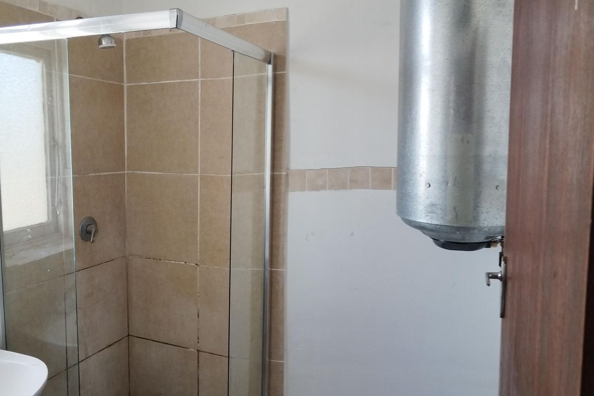 A functional bathroom featuring a glass-enclosed shower with tiled finishes, a wash basin, and exposed plumbing and water heater infrastructure.