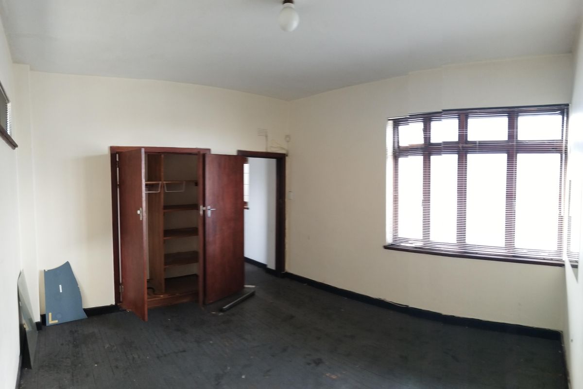 An empty bedroom featuring a built-in wooden cupboard, large window with blinds, and dark carpet flooring.