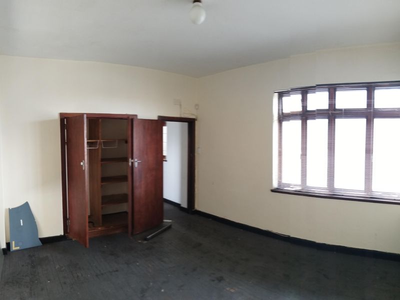 An empty bedroom featuring a built-in wooden cupboard, large window with blinds, and dark carpet flooring.