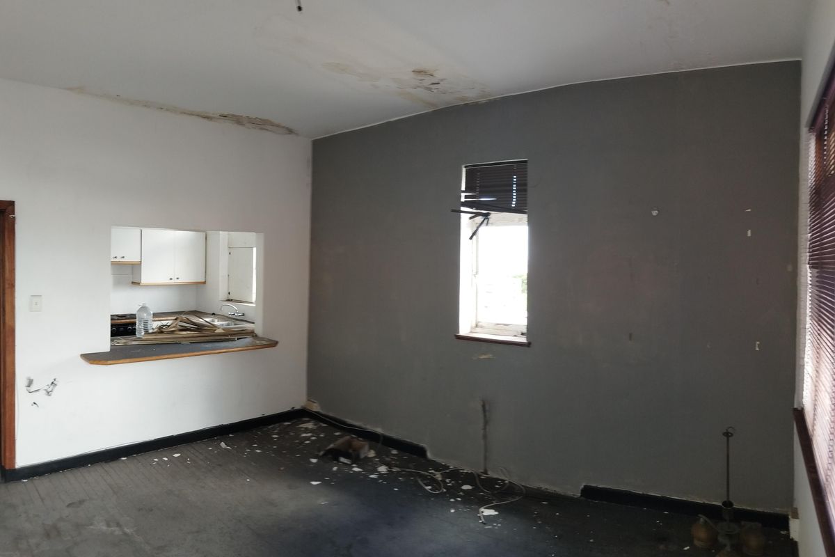 An empty living room area featuring a kitchen serving hatch, showing significant signs of wear including ceiling water damage and damaged flooring.