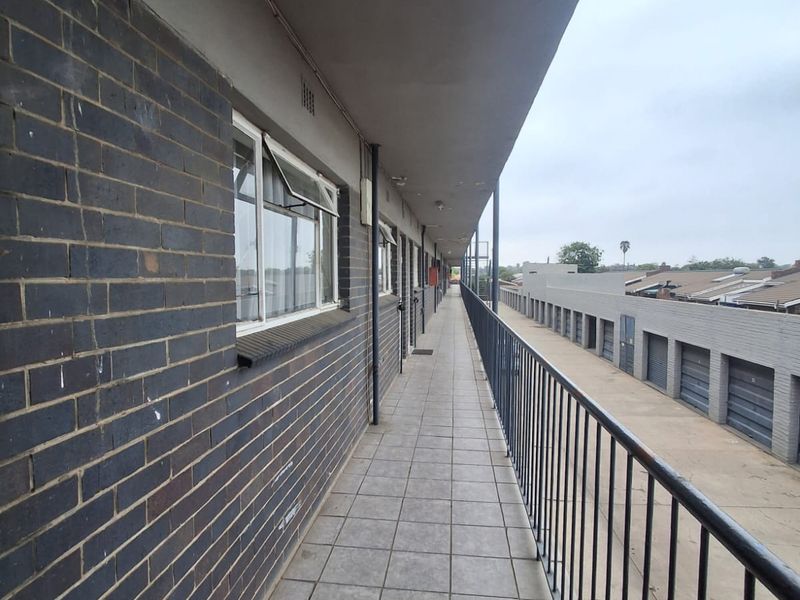 An exterior walkway or balcony featuring tiled flooring, a metal safety railing, and brick wall construction overlooking a garage complex.