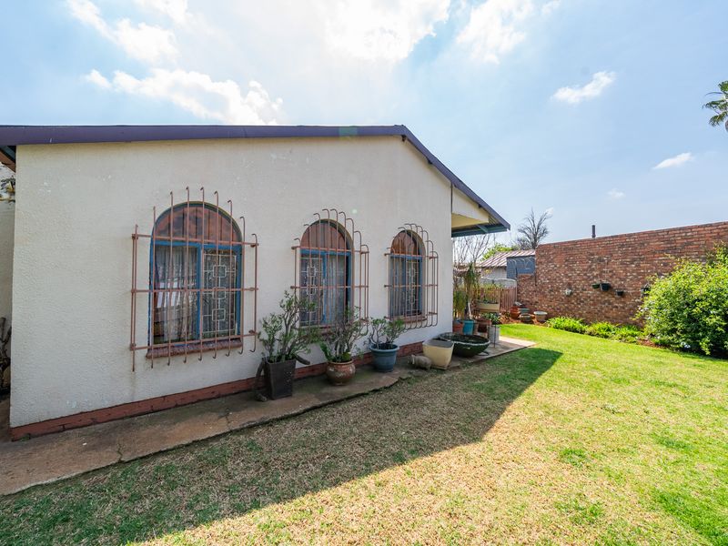 Rear view of the property featuring a lawn, concrete pathway, and arched windows with security bars set against a brick boundary wall.