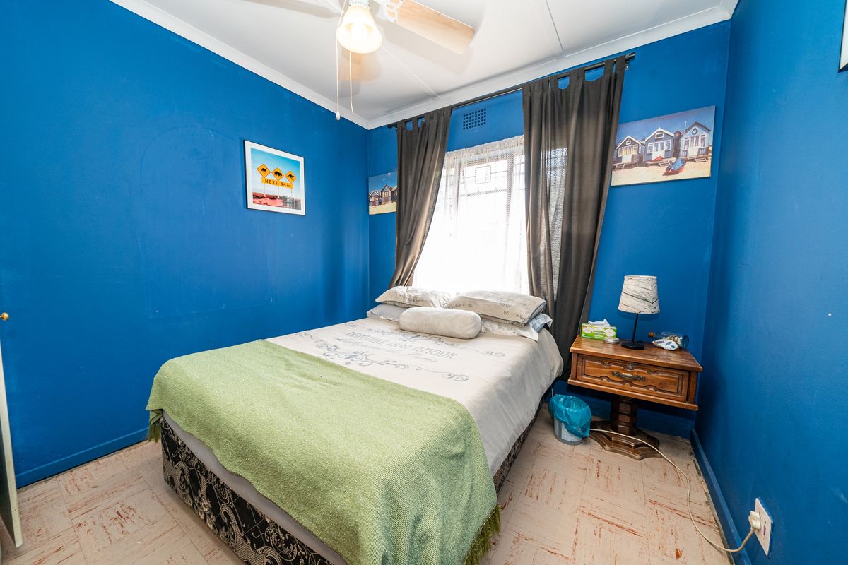 A bedroom featuring blue painted walls, a ceiling fan, and a window with curtains. The room has dated flooring and requires cosmetic updates.