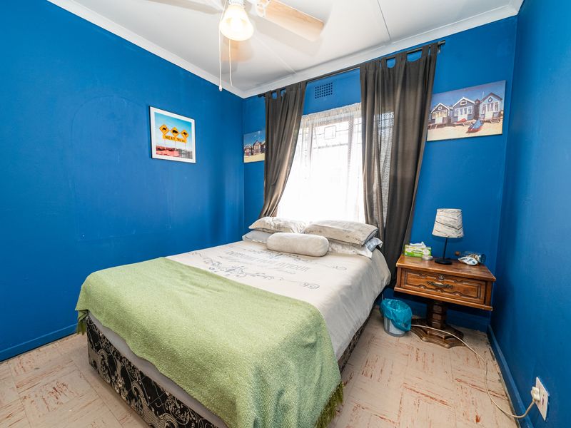 A bedroom featuring blue painted walls, a ceiling fan, and a window with curtains. The room has dated flooring and requires cosmetic updates.