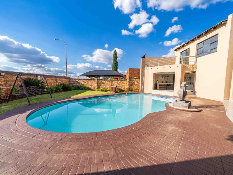 A well-maintained outdoor pool area featuring a curved swimming pool, brick paving, a water feature, and a rear garden with a brick boundary wall.