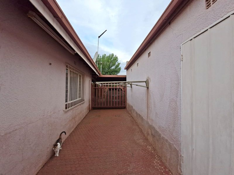 A narrow paved side passage between two exterior walls, featuring a security gate, window bars, and a wall-mounted clothesline.