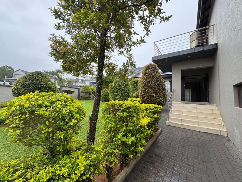 A well-maintained rear garden featuring a paved walkway, tiled staircase leading to the entrance, and an upper-level balcony with stainless steel railings.