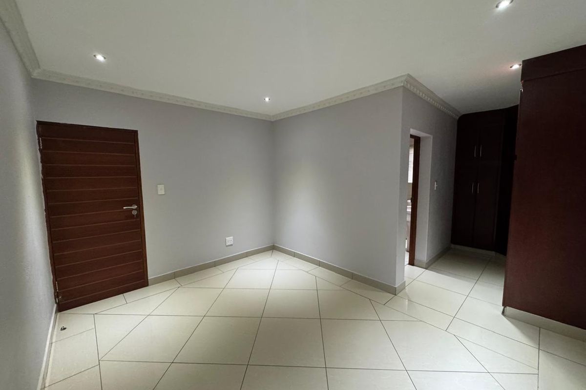 A clean, empty bedroom featuring neutral grey walls, tiled flooring, recessed downlighting, and built-in wooden cupboards.