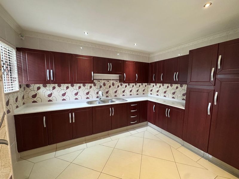 A spacious kitchen featuring dark wood-toned built-in cabinetry, white countertops, a stainless steel sink, and tiled flooring with a decorative backsplash.