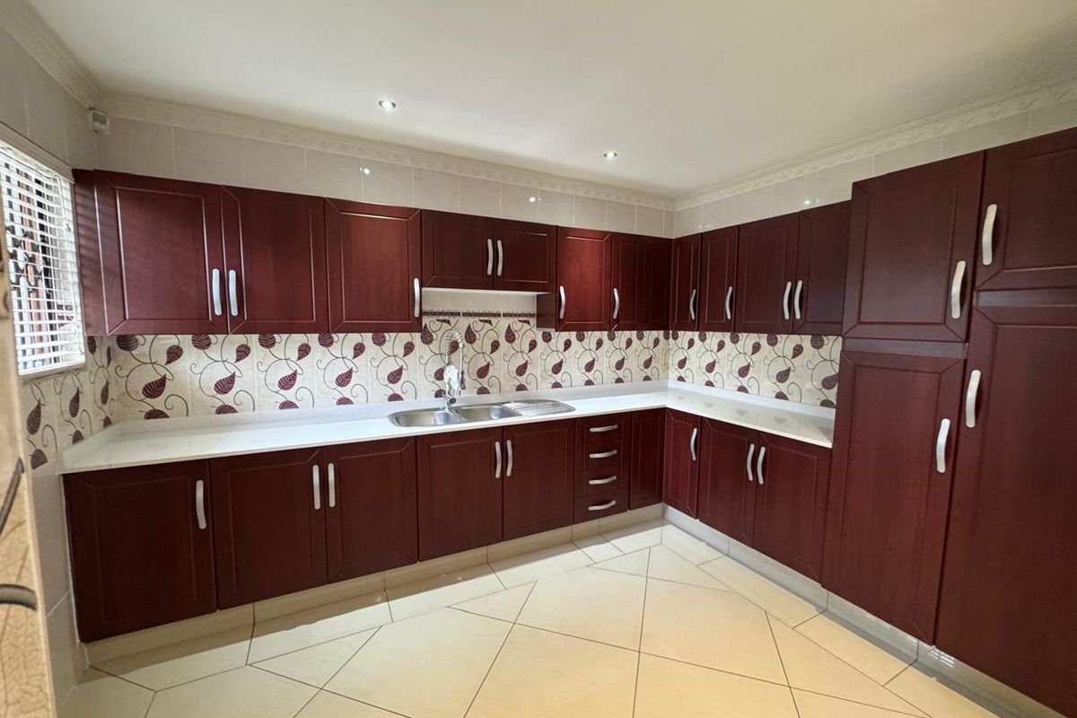 A spacious kitchen featuring dark wood-toned built-in cabinetry, white countertops, a stainless steel sink, and tiled flooring with a decorative backsplash.