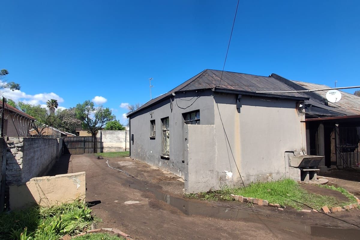 Rear exterior view of the property featuring a concrete boundary wall, metal roofing, and an external wash basin area with unpaved ground.
