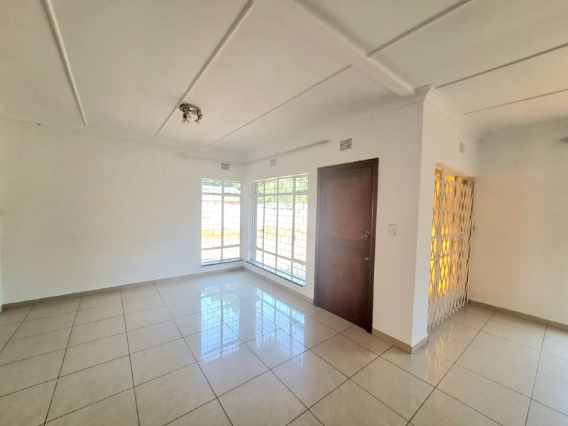 A bright, vacant living area featuring neutral tiled flooring, security gates, and large windows with burglar bars, offering a clean canvas for personalization.