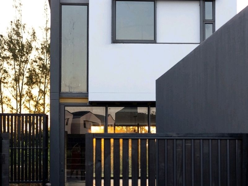 Modern townhouse exterior featuring a clean white and dark grey facade, large glass windows, and a private paved entrance with metal slat fencing.