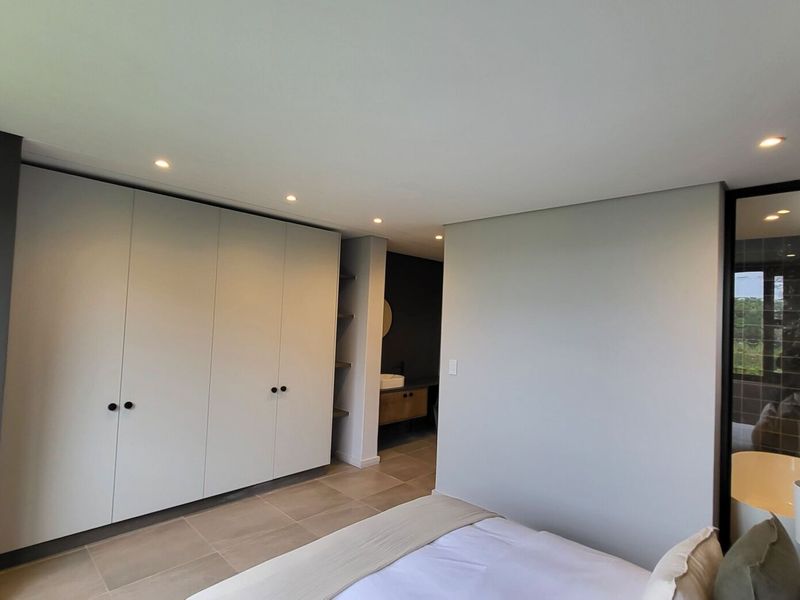 Modern bedroom featuring built-in cupboards, recessed downlighting, and tiled flooring, with a seamless transition to an en-suite bathroom area.
