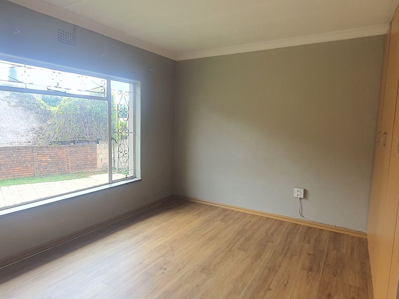 A bright, empty bedroom featuring laminate flooring, built-in cupboards, and a large window with security bars overlooking the garden.