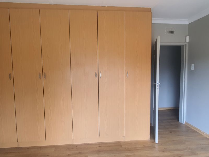A spacious bedroom featuring light wood-finish built-in cupboards, laminate flooring, and neutral grey walls, offering a clean and functional living space.