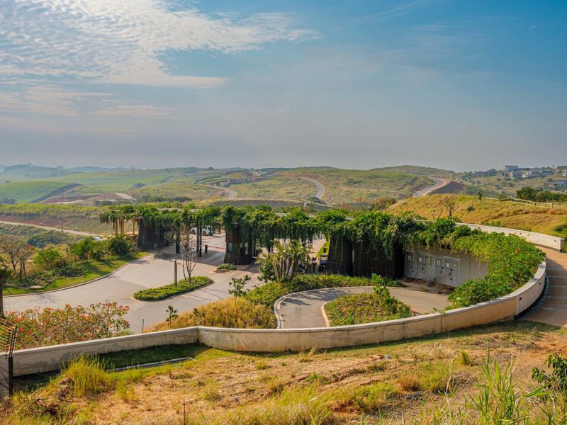 A high-angle view of a modern, landscaped gated estate entrance featuring a curved retaining wall and integrated security gatehouse structure.