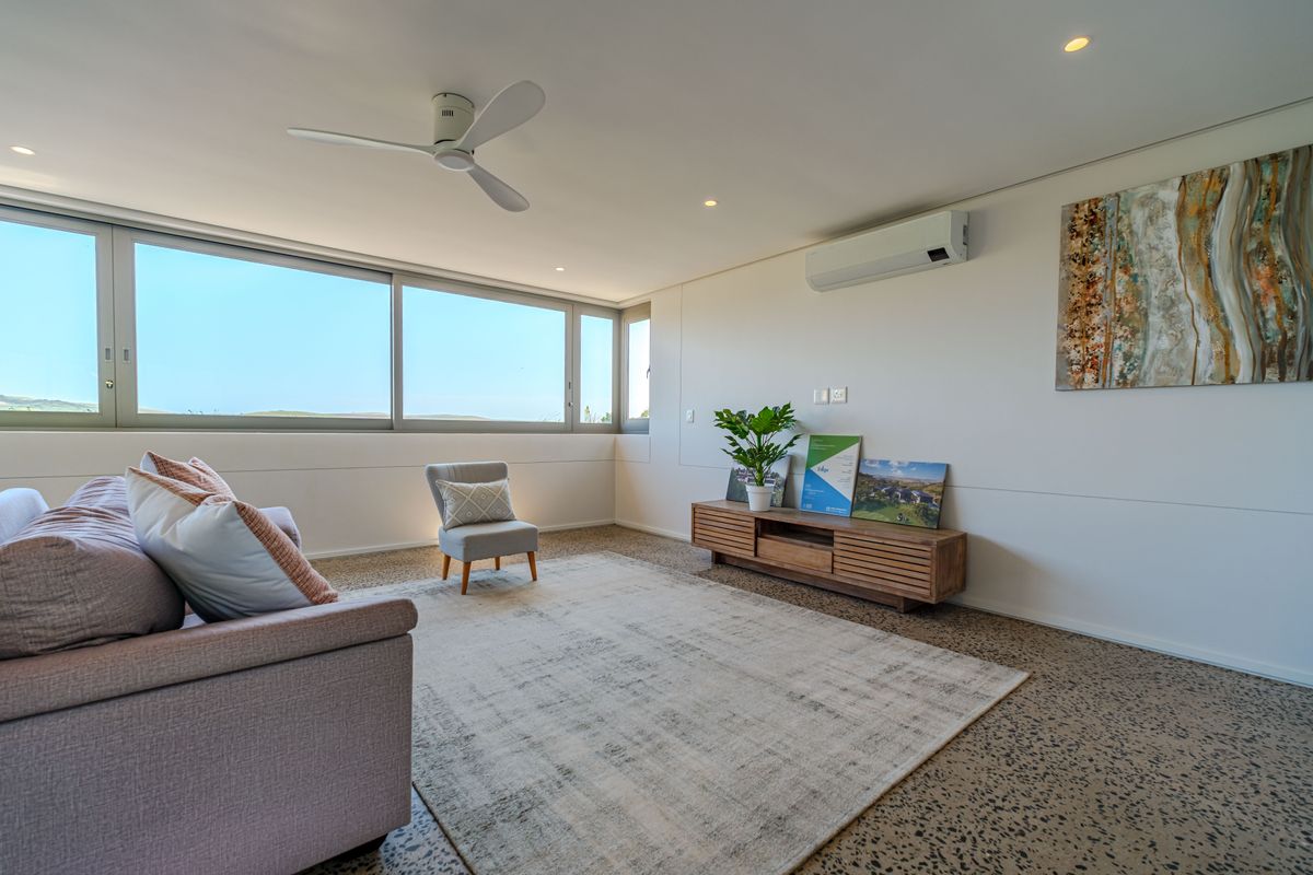 A bright and modern living room featuring polished concrete floors, a ceiling fan, split-system air conditioning, and large windows offering natural light.