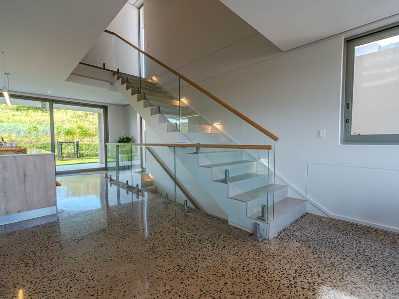 Modern open-plan living area featuring a striking floating staircase with glass balustrades, polished concrete floors, and large sliding doors leading to the garden.