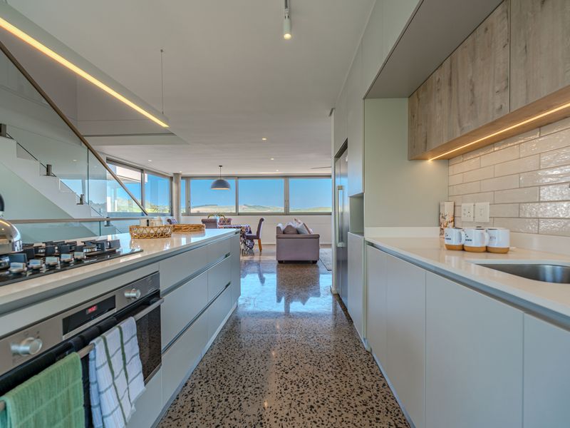 Modern open-plan kitchen featuring sleek cabinetry, stone countertops, gas cooktop, and polished concrete floors, flowing into a bright living area with scenic views.