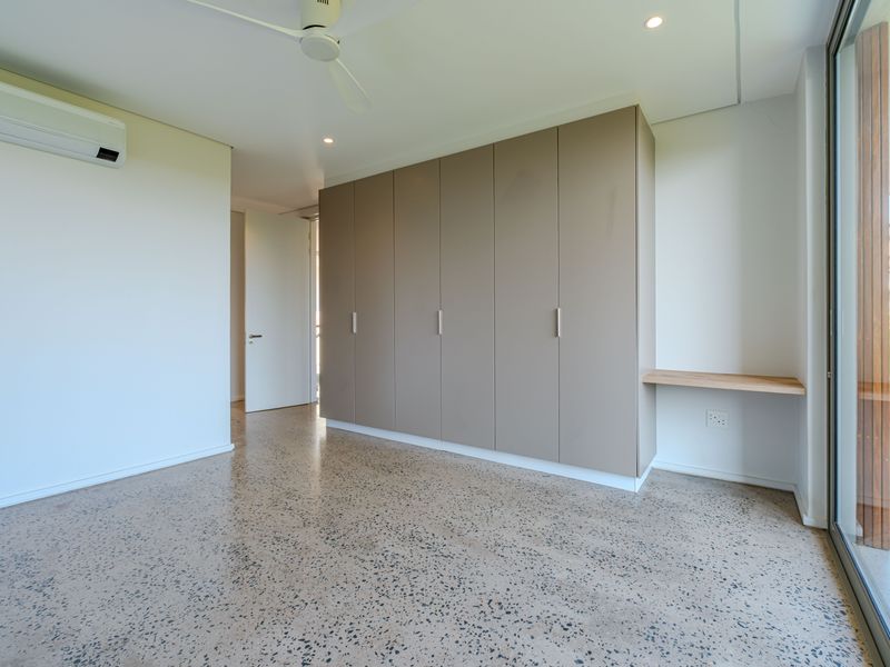 A bright, modern bedroom featuring polished concrete floors, extensive built-in cabinetry, a floating desk, and direct access to an outdoor balcony.