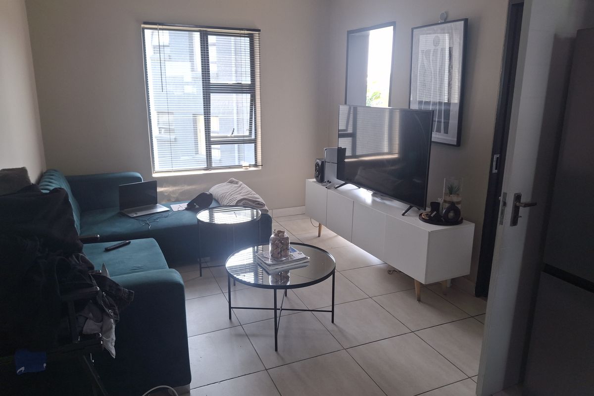 A functional living room featuring neutral tiled flooring and a large window with venetian blinds, providing natural light to the space.