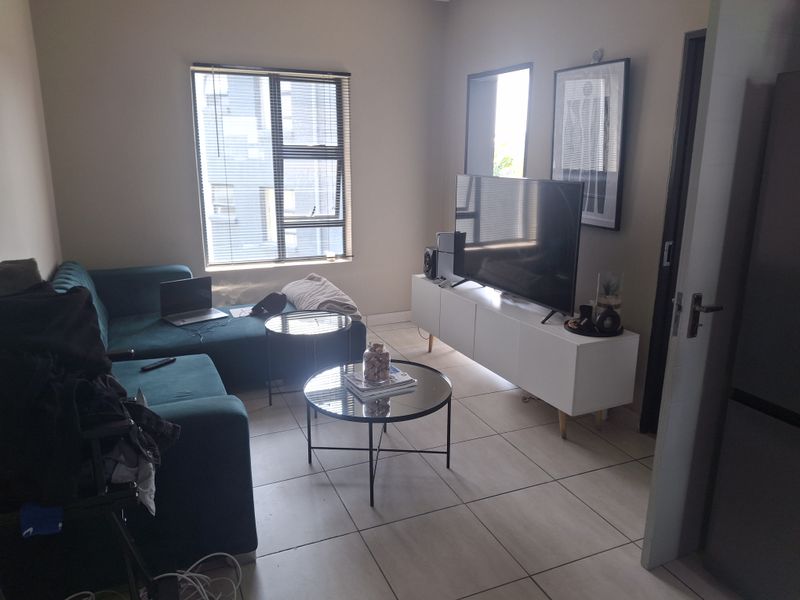 A functional living room featuring neutral tiled flooring and a large window with venetian blinds, providing natural light to the space.