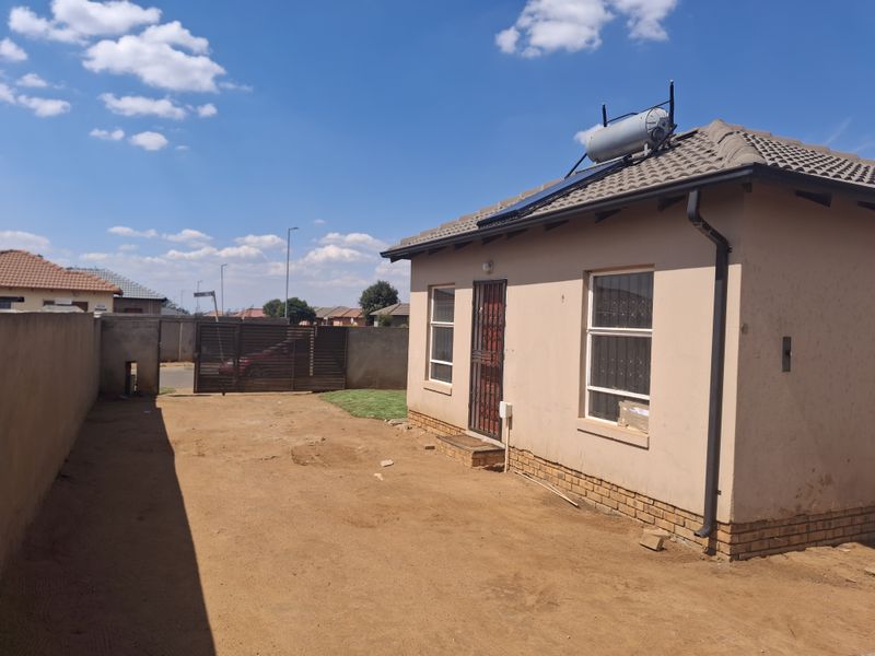 Rear view of the property featuring a tiled roof with a solar geyser, security gate, and a large unpaved yard area enclosed by perimeter walls.
