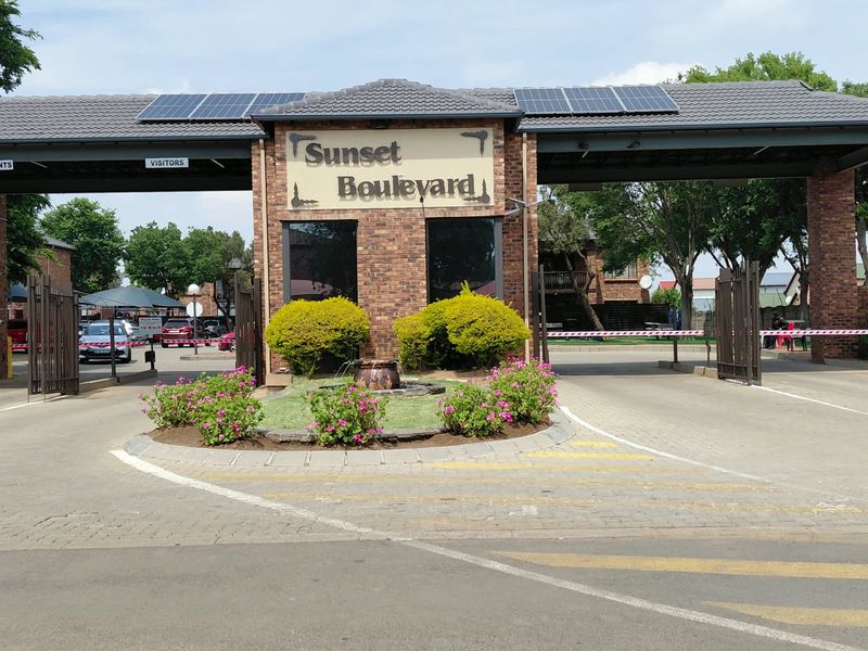 The main entrance and security gatehouse of the Sunset Boulevard townhouse complex, featuring brick construction, solar panels, and a landscaped garden.