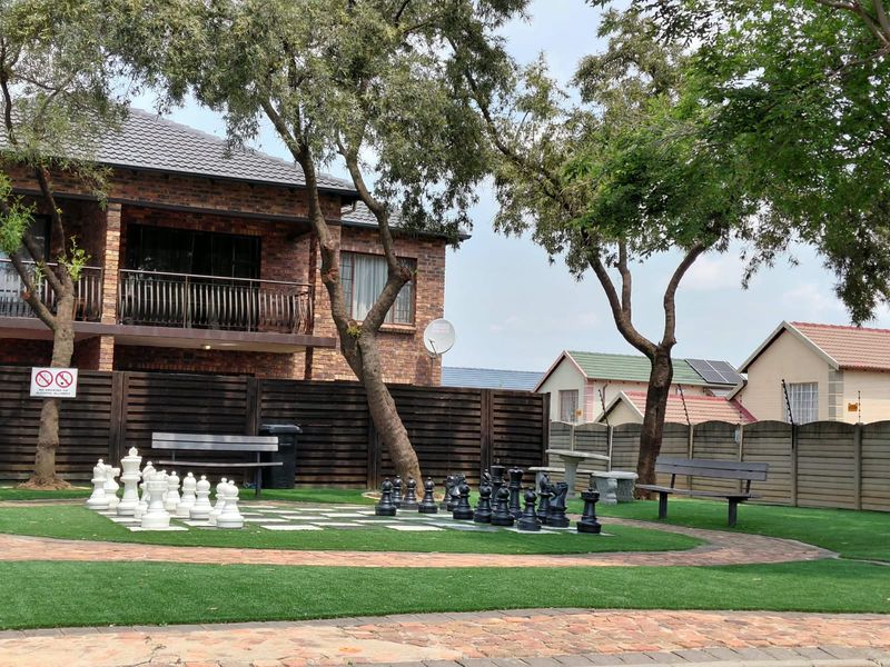 A landscaped communal garden area featuring a large outdoor chess set, artificial lawn, paved pathways, and perimeter fencing within a townhouse complex.