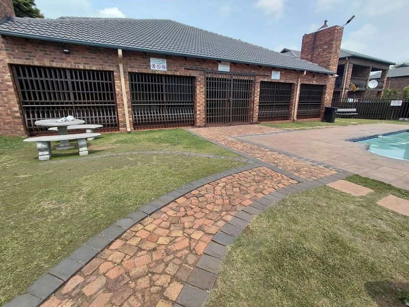 Communal pool area featuring a brick-built clubhouse with security gates, paved pathways, and landscaped lawn space.