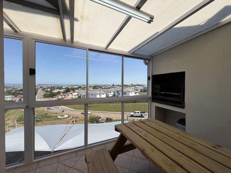Enclosed balcony featuring a built-in braai, tiled flooring, and expansive windows offering panoramic coastal and neighbourhood views.