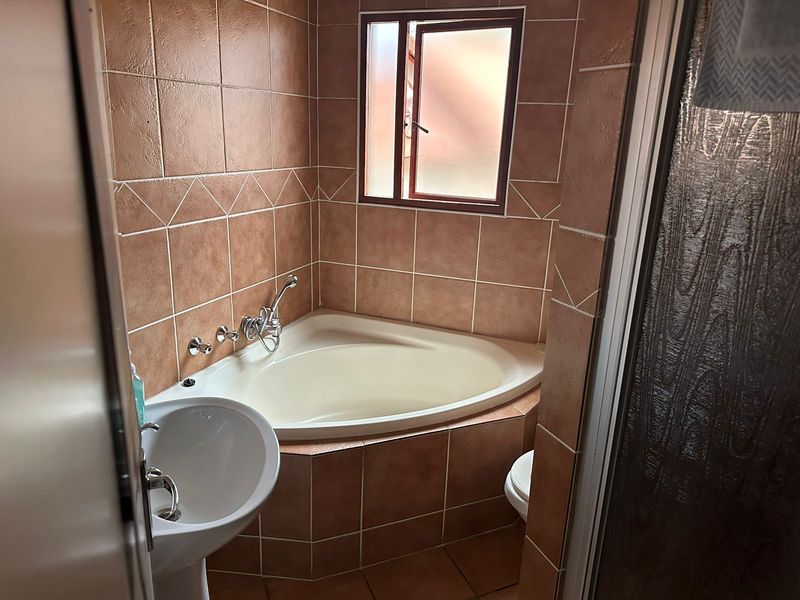A compact bathroom featuring a corner bathtub, pedestal basin, and floor-to-ceiling tiled finishes with a small casement window for natural light.