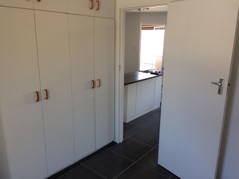 A clean hallway featuring extensive built-in storage cupboards and dark floor tiles, leading through to an open-plan kitchen area.