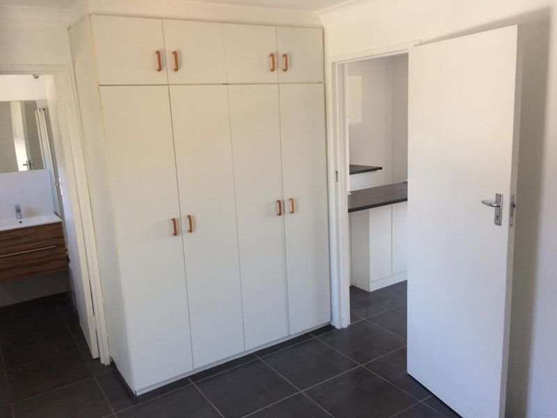 A clean, empty bedroom featuring built-in cupboards, tiled flooring, and direct access to an adjoining bathroom and hallway.