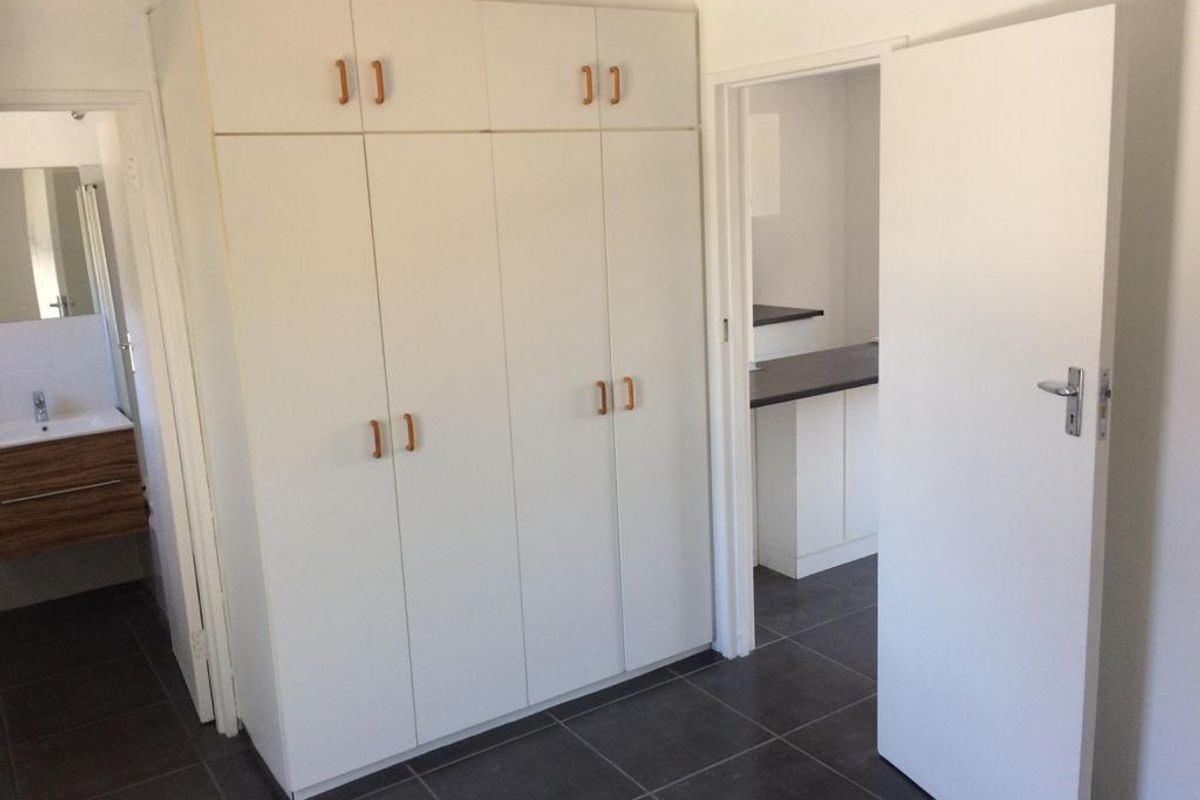 A clean, empty bedroom featuring built-in cupboards, tiled flooring, and direct access to an adjoining bathroom and hallway.