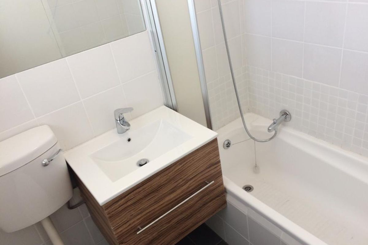 A clean, modern bathroom featuring a wall-mounted wood-grain vanity, white basin, toilet, and a combined bathtub and shower unit with white wall tiling.
