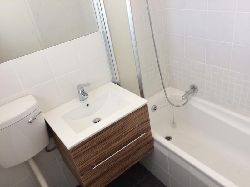 A clean, modern bathroom featuring a wall-mounted wood-grain vanity, white basin, toilet, and a combined bathtub and shower unit with white wall tiling.