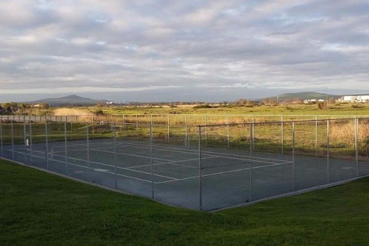 Outdoor hard-court tennis facility enclosed by chain-link fencing, situated in an open landscape setting.