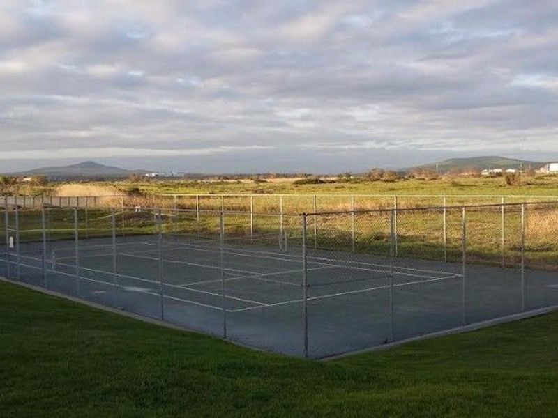 Outdoor hard-court tennis facility enclosed by chain-link fencing, situated in an open landscape setting.
