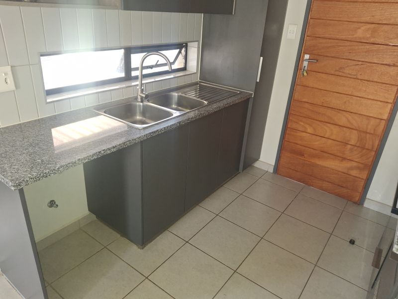 A clean and modern kitchen area featuring granite countertops, a stainless steel double sink, tiled backsplash, and neutral floor tiles.