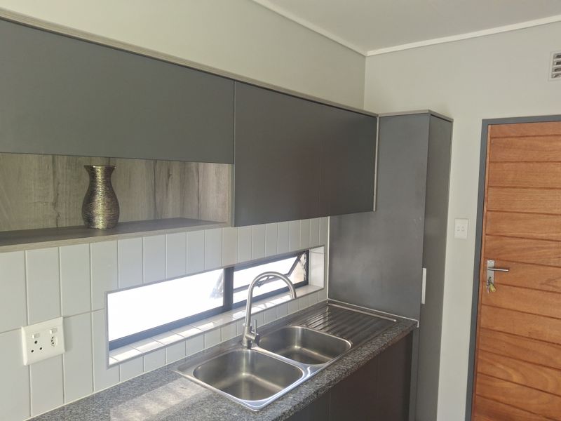 Modern kitchen area featuring sleek grey cabinetry, granite countertops, a double stainless steel sink, and a tiled backsplash with a horizontal window.