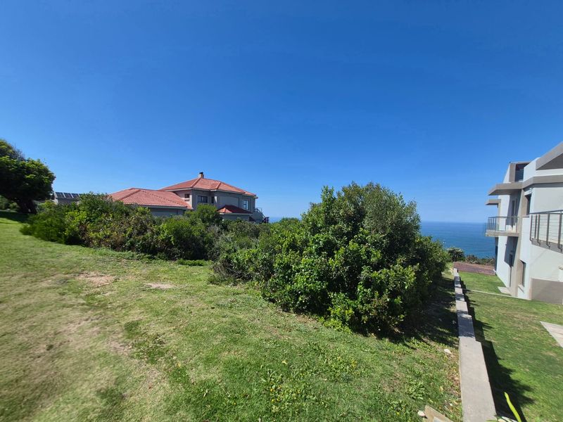A well-maintained residential garden area featuring a retaining wall, mature shrubs, and expansive lawn space with views of the surrounding coastal landscape.