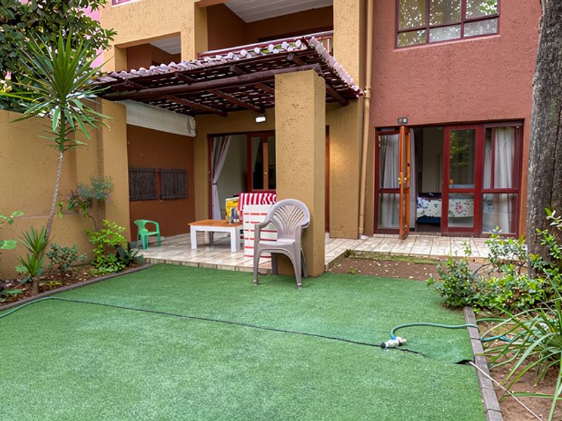 Rear exterior view of the apartment featuring a covered patio with timber pergola, tiled flooring, and a private garden area with artificial lawn.