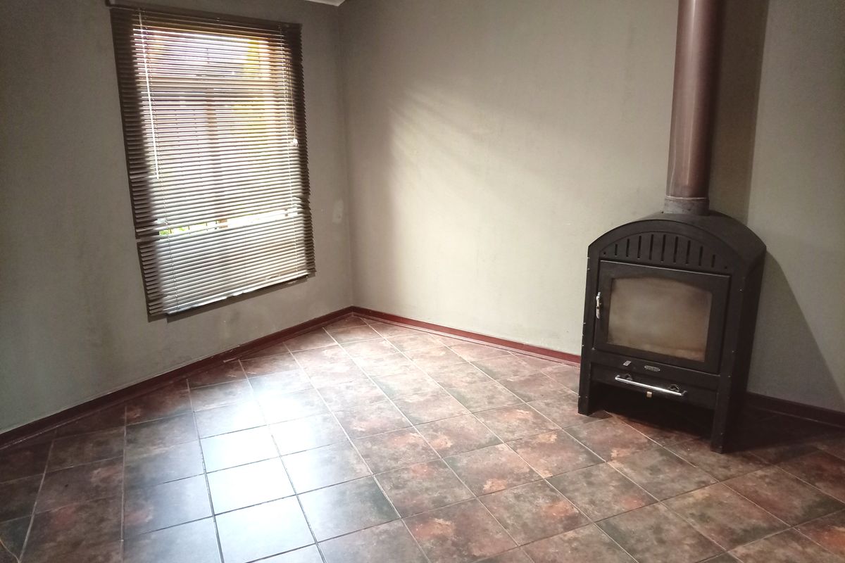An empty living room featuring tiled flooring, a freestanding fireplace with a flue, and venetian blinds on the window.