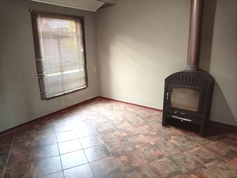 An empty living room featuring tiled flooring, a freestanding fireplace with a flue, and venetian blinds on the window.