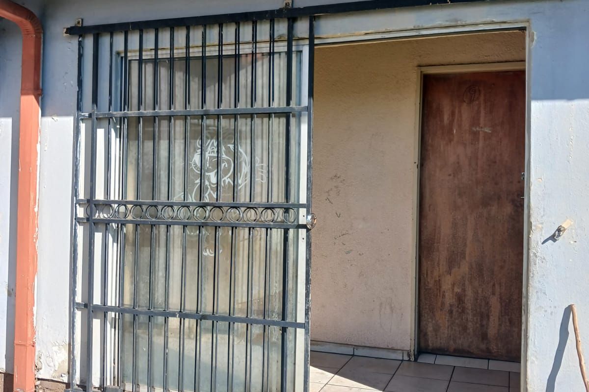 Exterior view of a rear entrance featuring a sliding security gate, tiled threshold, and weathered wall finishes requiring maintenance.