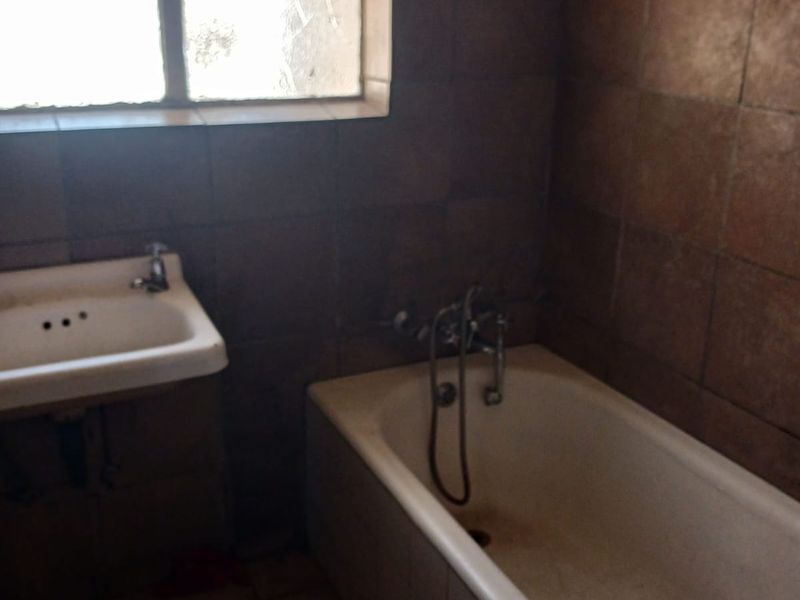 A dated bathroom featuring a built-in bathtub, wall-mounted basin, and dark tiled finishes, requiring modernization to improve its overall condition and appeal.