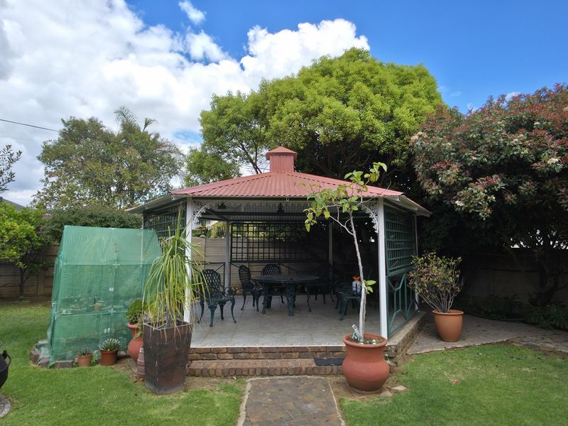 A covered outdoor gazebo featuring a metal roof, tiled flooring, and brick foundation, set within a landscaped garden area.